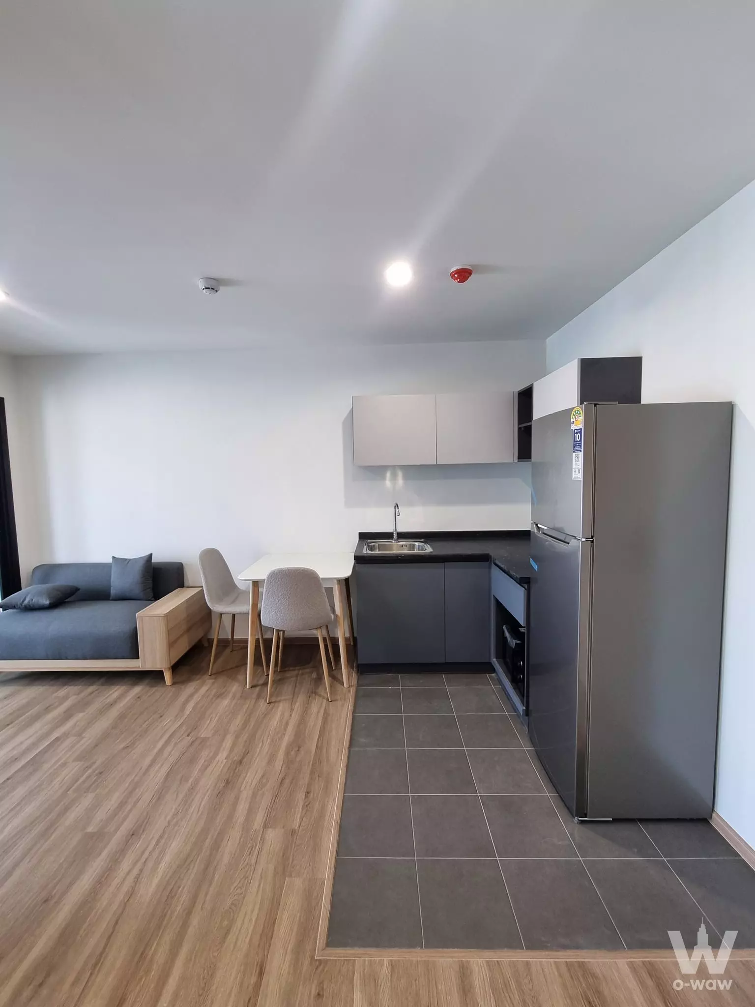 Open-plan living area in ARLO Sukhumvit 105 with grey sofa, dining set, and L-shaped kitchenette cabinetry