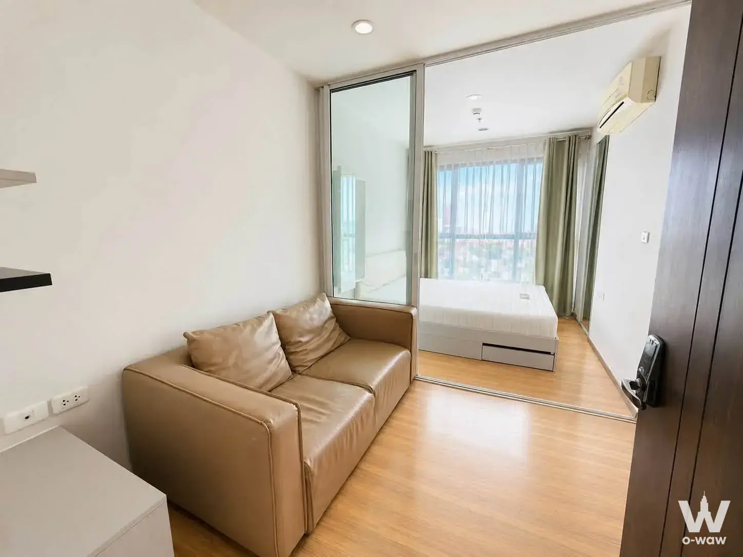Open-plan studio condo for sale in THE BASE Chaengwattana, featuring tan leather seating, wood floors, and a glass partition