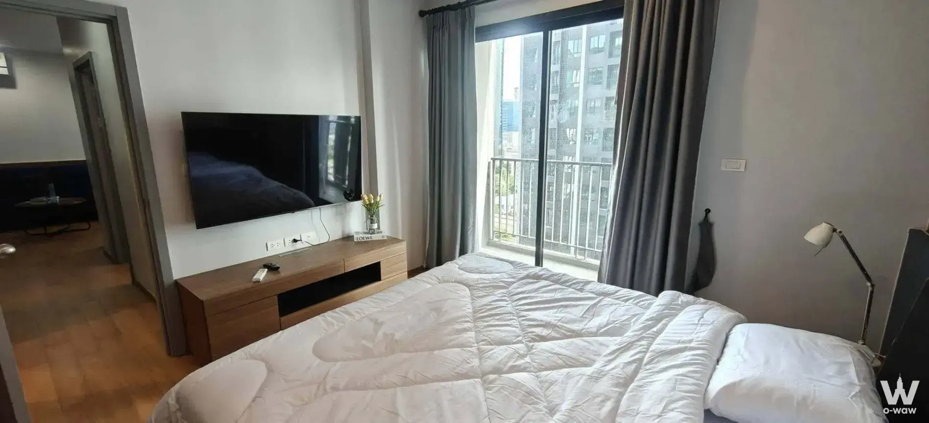 Bedroom with queen bed, wall-mounted TV, and sliding glass doors leading to a balcony overlooking adjacent high-rises in a