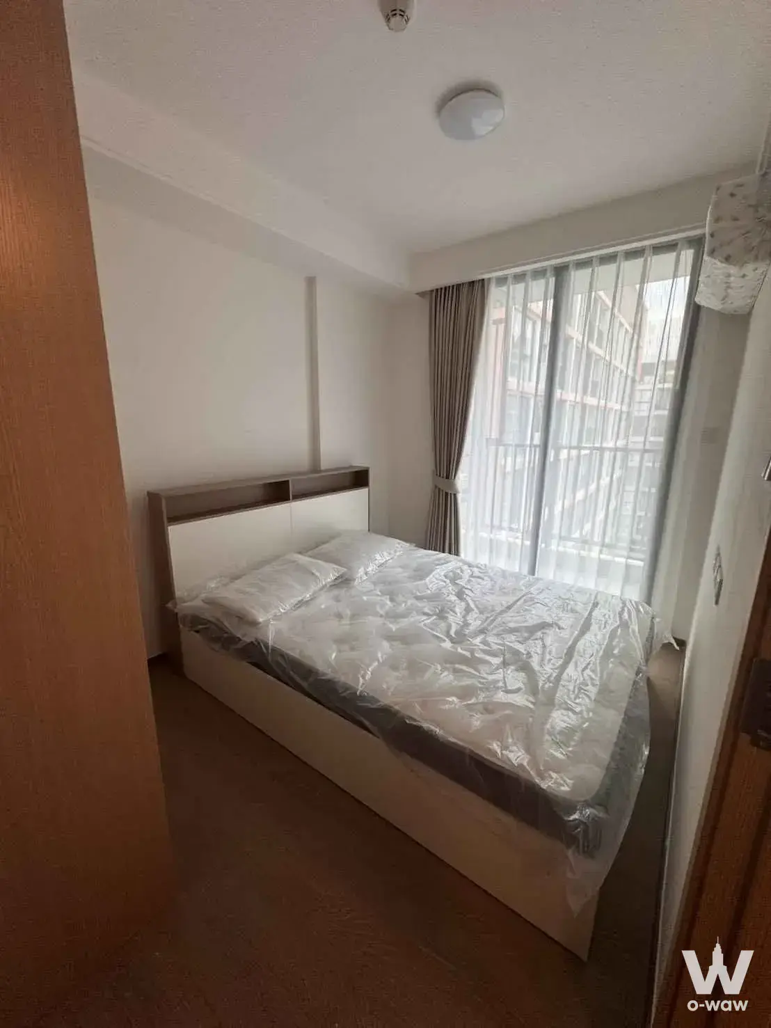 Small bedroom in Regal Srinakarin 40 featuring a single bed with protective wrap, dark brown laminate flooring, and balcony