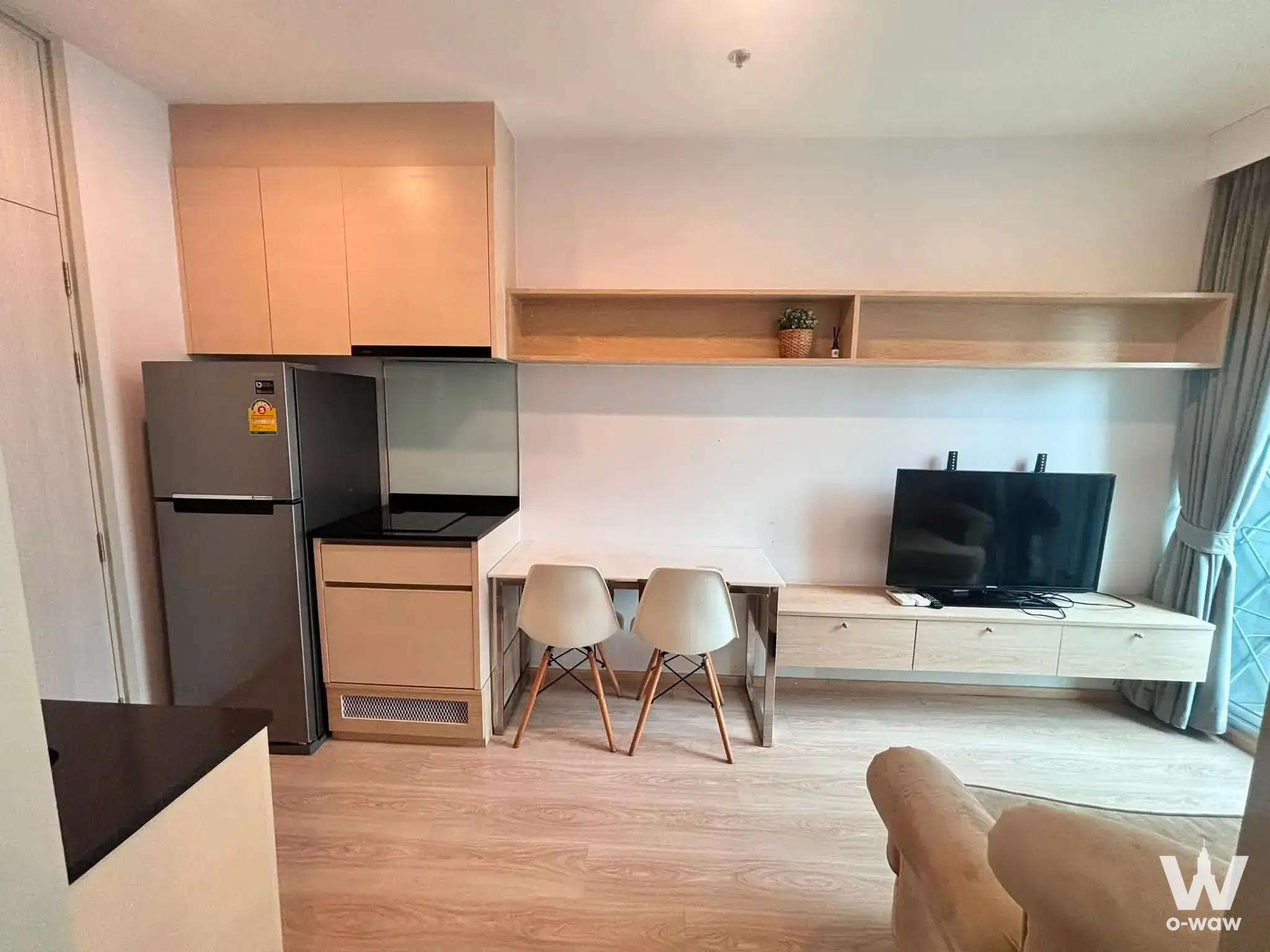 Open-plan living area with light wood floors, a black glass cooktop, and a small desk in this Ratchada 2 condo