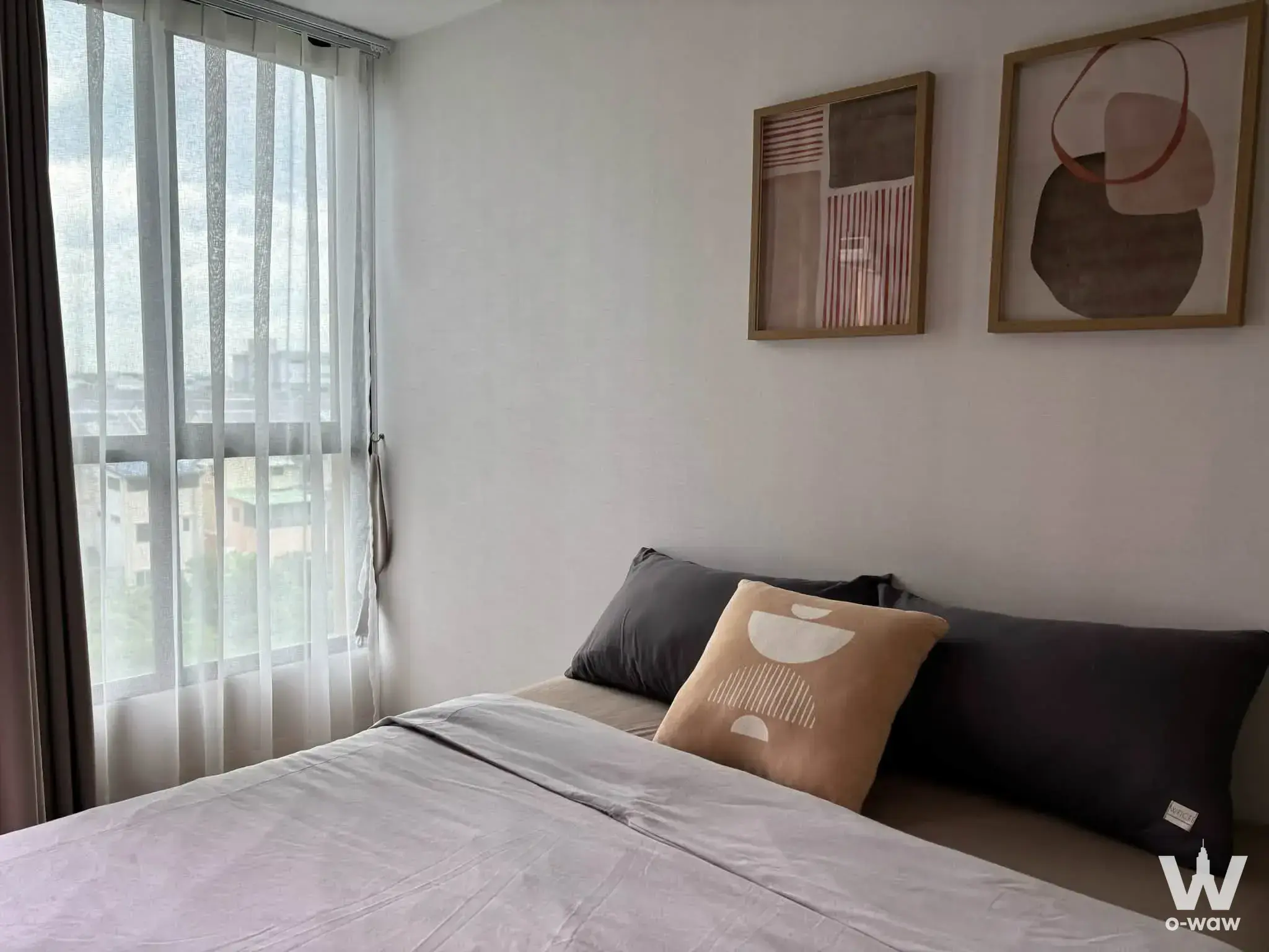 The bedroom in a 1-bed Atmoz Ratchada condo for rent has grey bedding, framed art and a city-view window