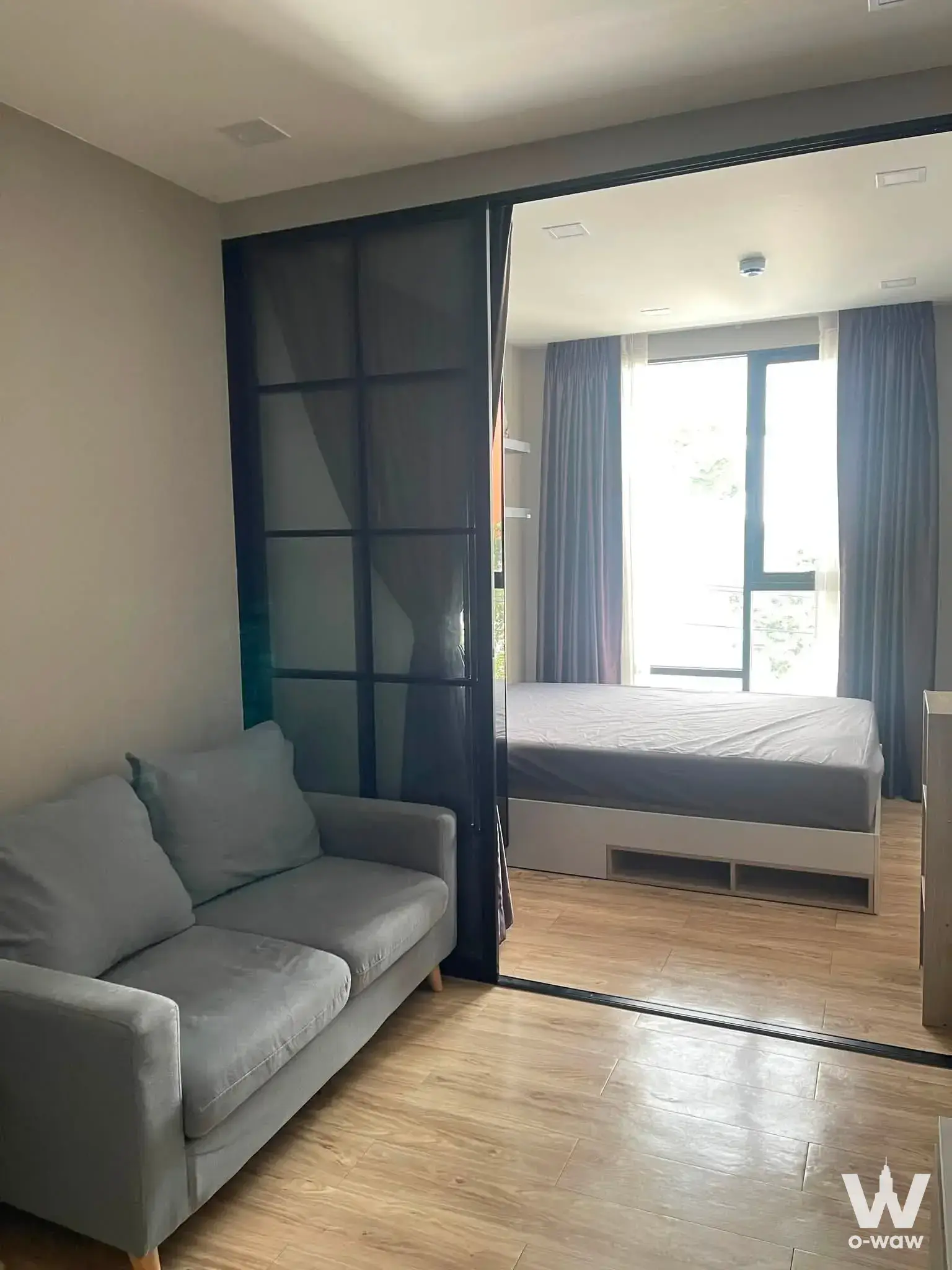 Open-plan studio with grey sofa and glass partition separating the bed area at Groove Ratchada-Ladprao
