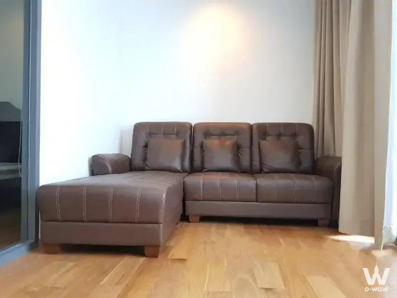 Living room in Hyde Sukhumvit 13 featuring brown leather sectional sofa on light wood floors with beige curtains