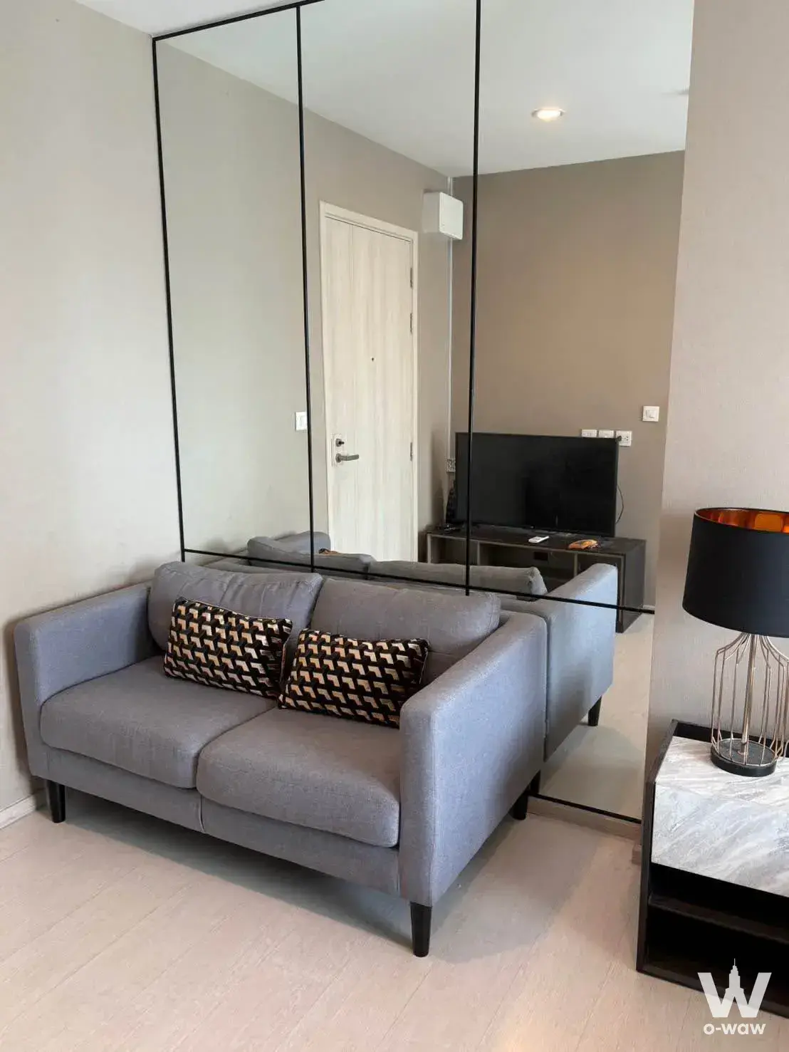 Modern living room in a Life Asoke 1-bed condo for rent, highlighting grey sofa, large mirror wall, and minimalist decor