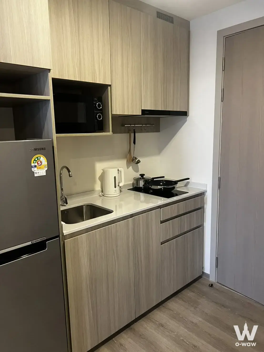 Compact kitchen with light wood cabinets, stainless steel fridge, and induction cooktop in a 1-bed condo for rent at Origin