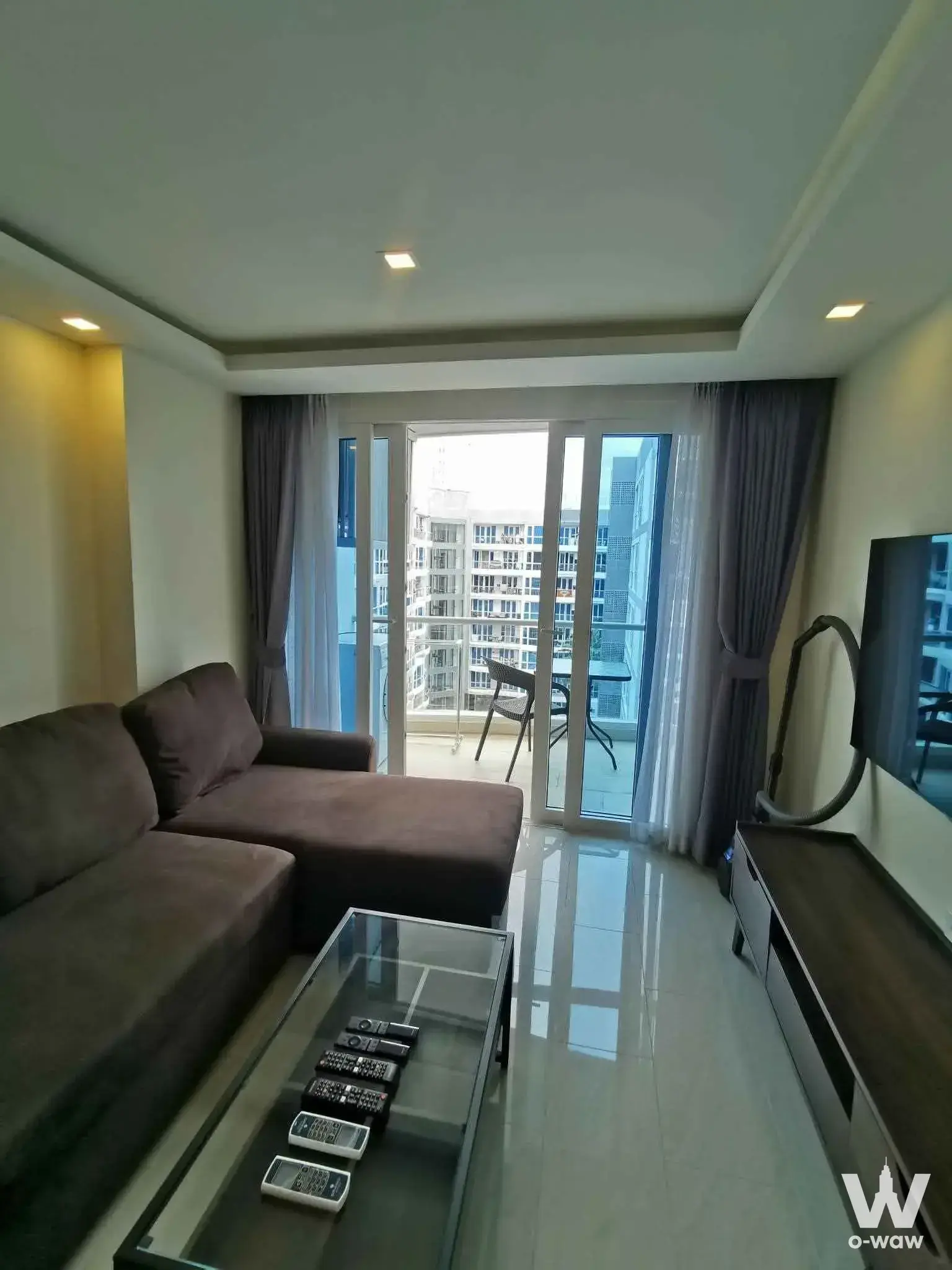 Living room has a dark-brown sectional, glass coffee table and balcony view in a 1-bed Grand Avenue Residence condo for rent