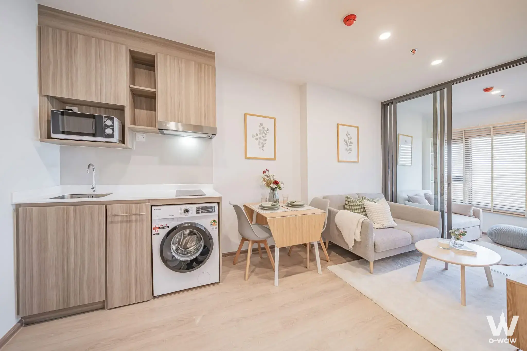 Open-plan kitchen and living area in Life Rama 4 Asoke condo featuring light wood cabinets and grey sofa