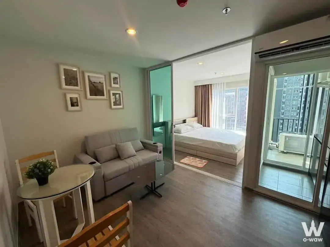 Open-plan studio in Regent Home Bangna with grey sofa, glass partition, and high-rise city views