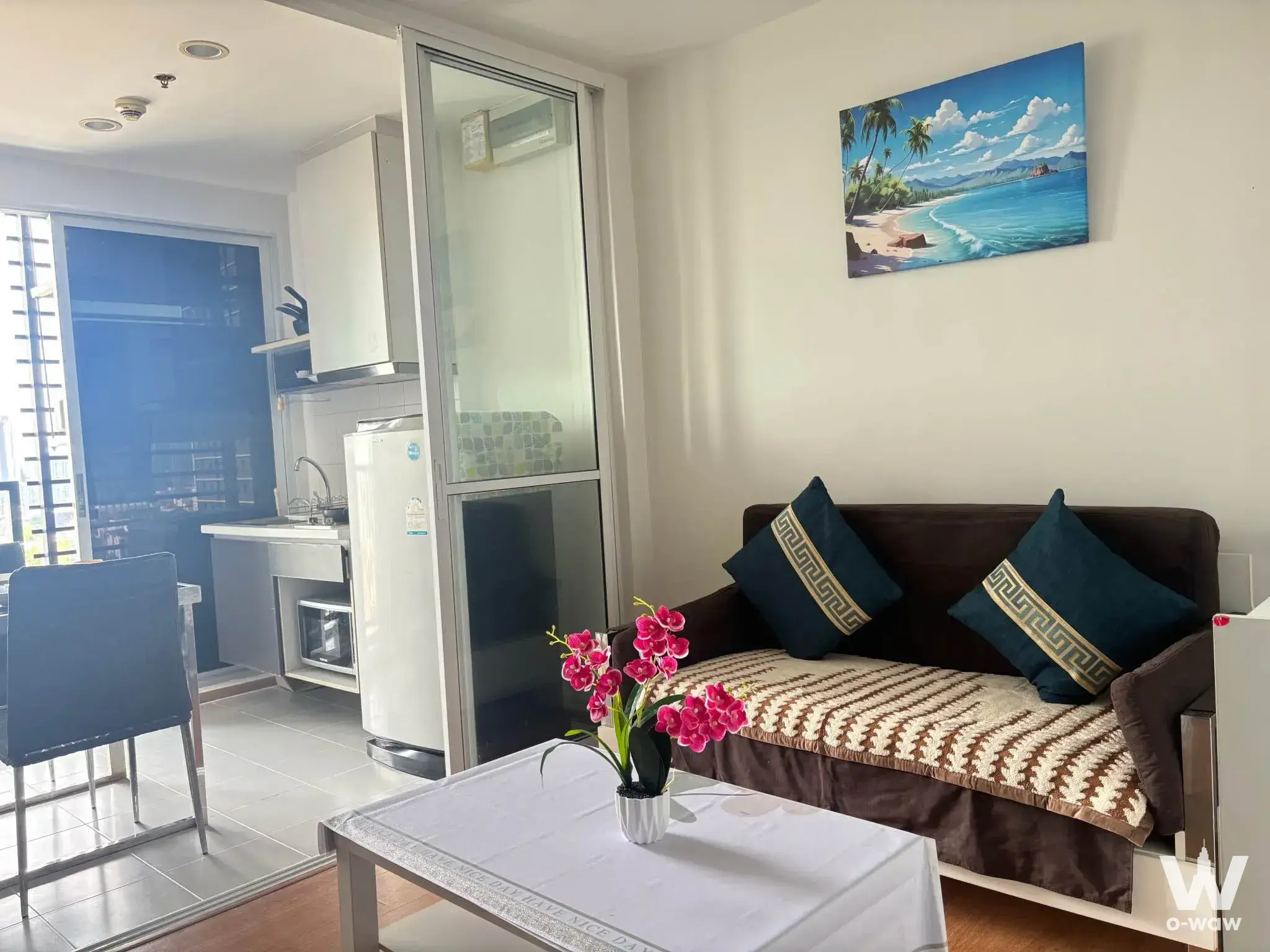 1-bed condo for sale in THE BASE Sukhumvit 77 living area with teal pillows, beach painting, and glass kitchen partition