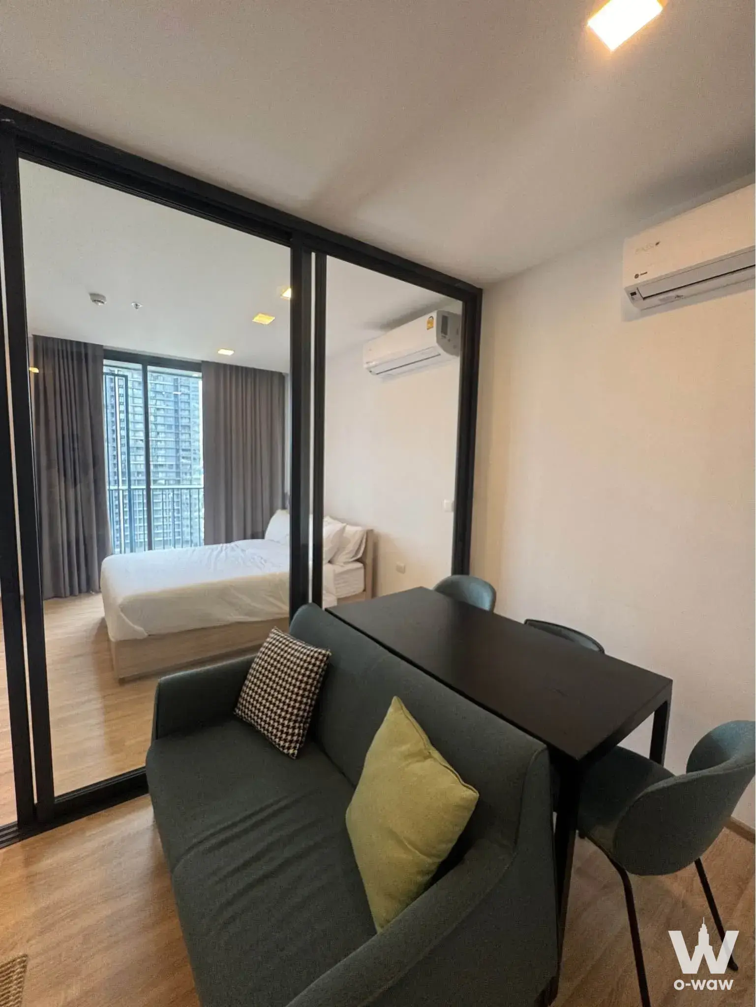 Open-plan condo at XT Phayathai with grey sofa, dining set, and glass partition to bedroom