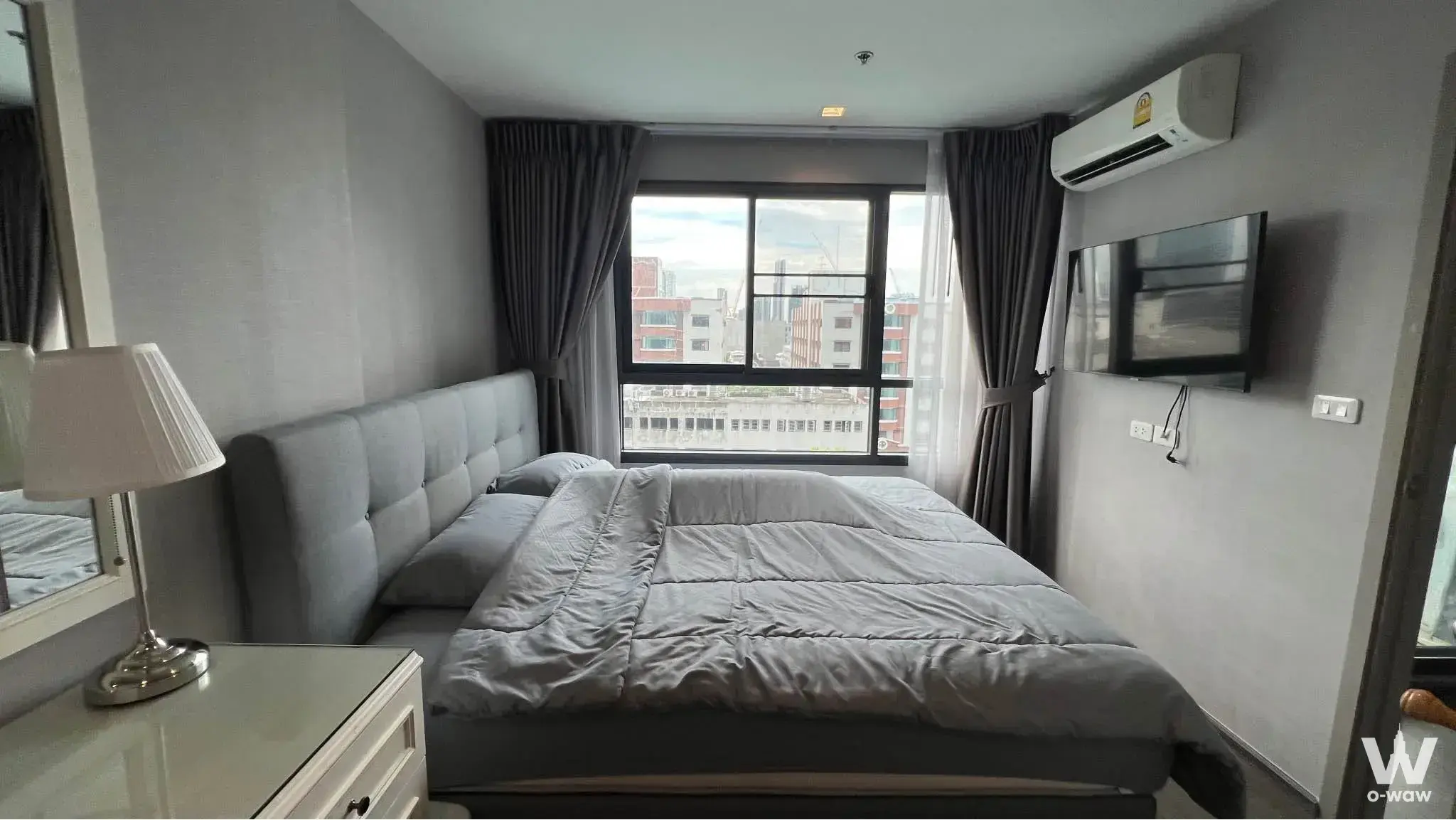 The bedroom in a 1-bed Ideo Sukhumvit 93 condo has a grey queen bed and a large city-view window