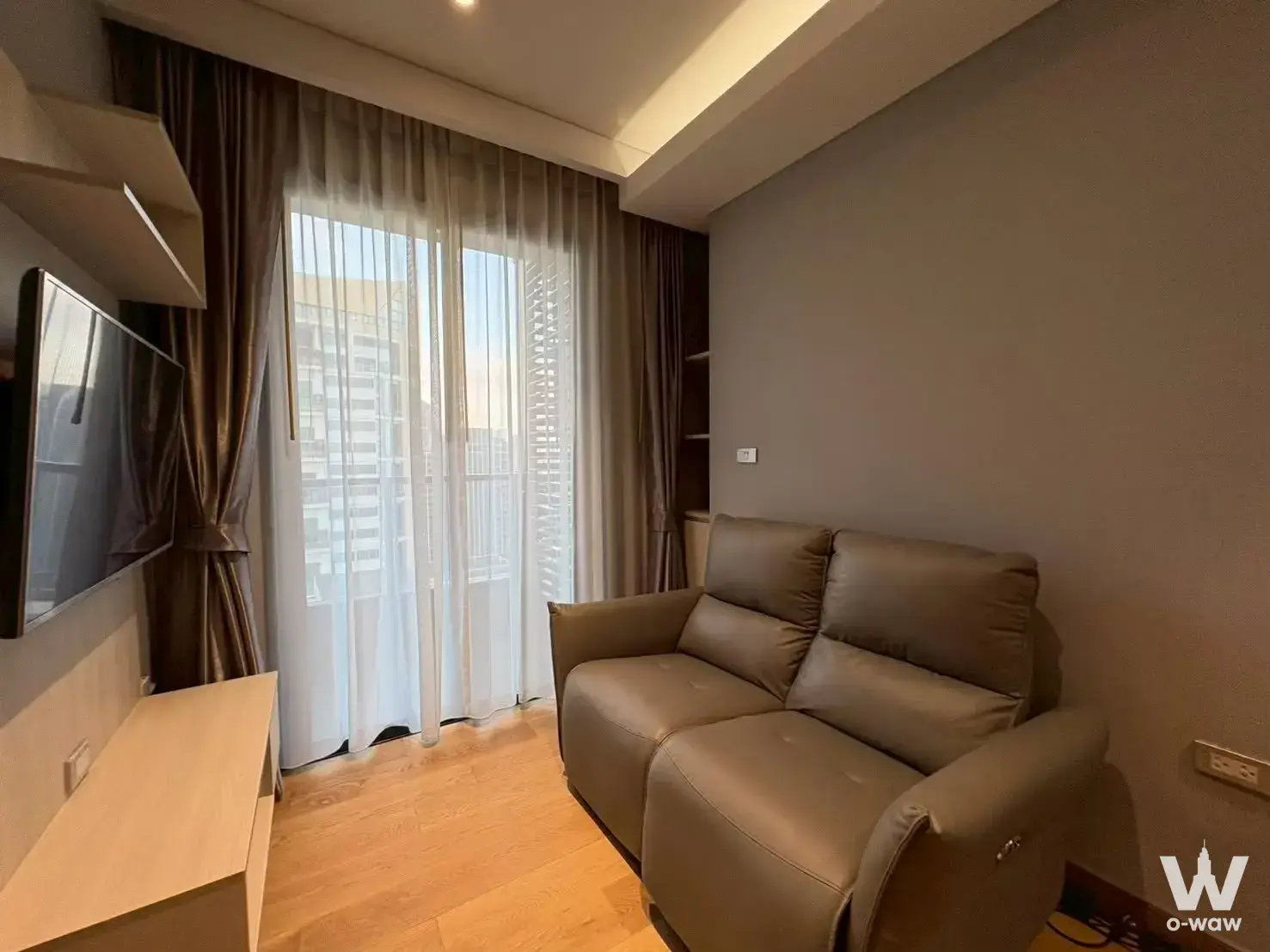 Living room in The Lumpini 24 featuring a taupe recliner and city view through glass doors