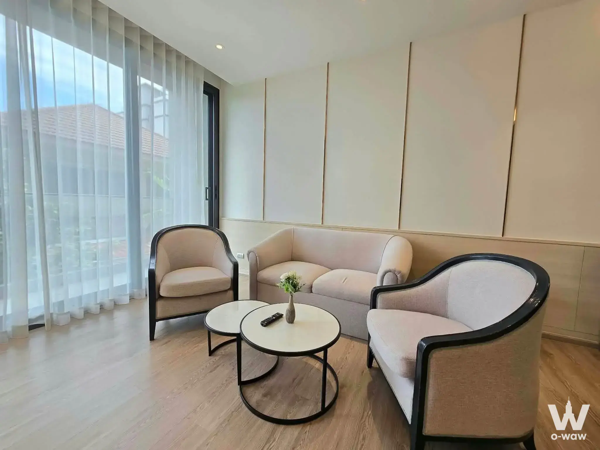 Bearing Residence living room with beige sofa, nested coffee tables, cream feature wall, and view of green trees