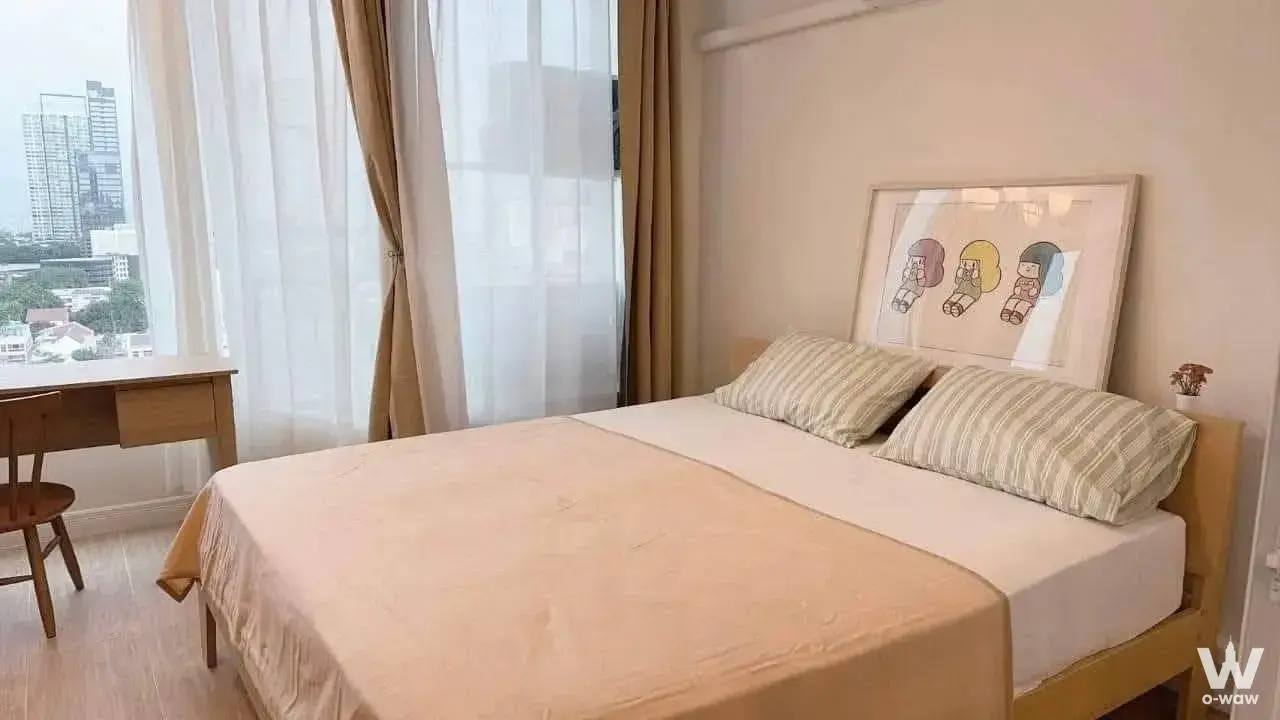 Queen bedroom in a La Maison 24 unit with peach bedspread, matching desk, and high-floor view of Bangkok buildings
