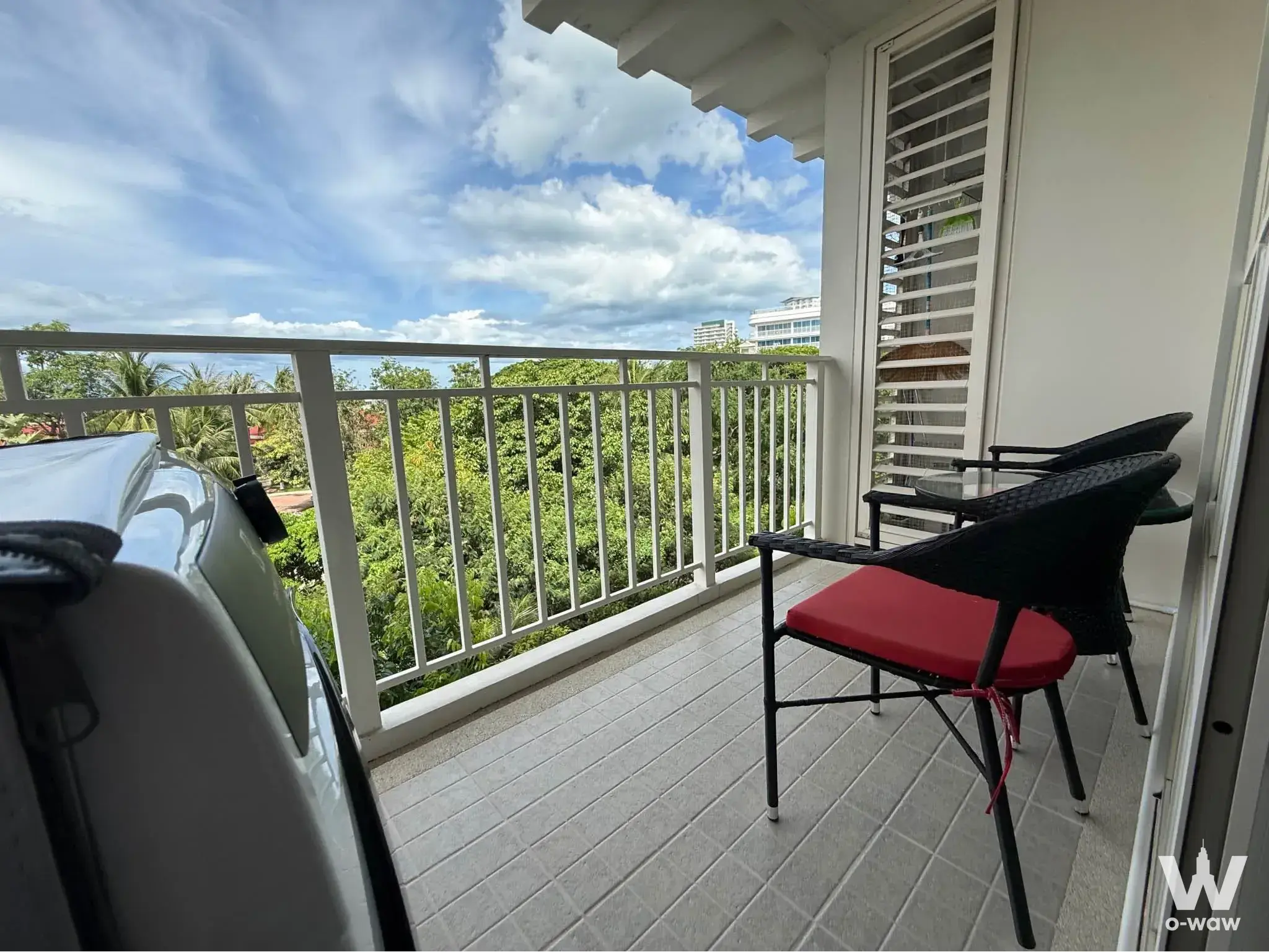 The balcony of a Baan Haad 1-bed rental condo has wicker chairs, a glass table and palm tree views