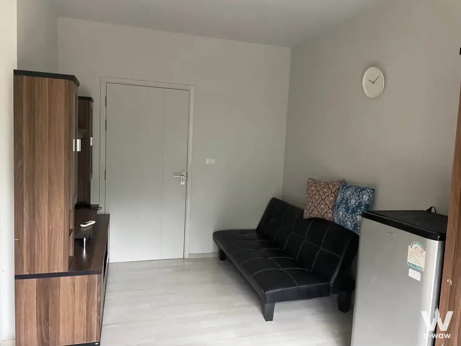 Compact living area with a black futuna sofa, dark wood entertainment cabinet, and mini-fridge in a studio for sale at UNiO