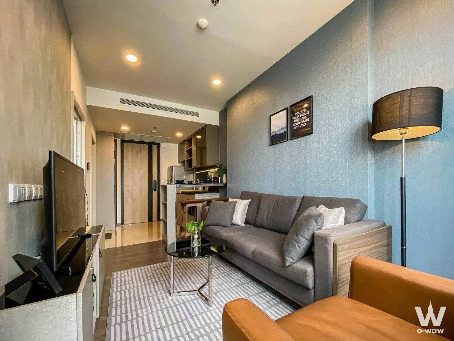 1-bed condo for rent in Oka Haus featuring an open-plan living area with a grey sofa and textured blue-grey accent wall