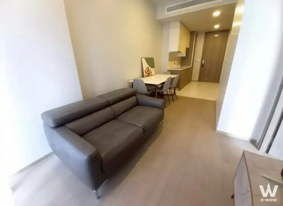 Open-plan living area in a Celes Asoke 1-bed condo with a dark leather sofa, dining table, and natural light