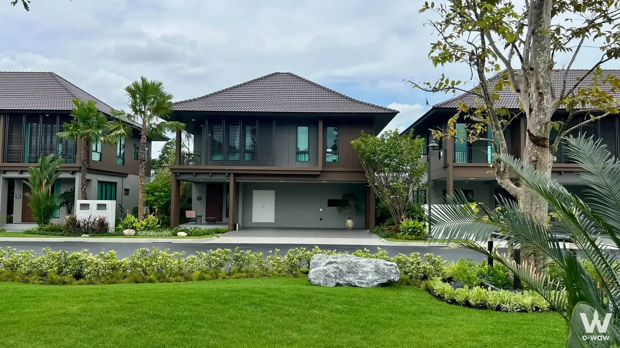 Two-story house for sale in Burasiri Well Bangkok with dark-brown siding and tropical landscaping