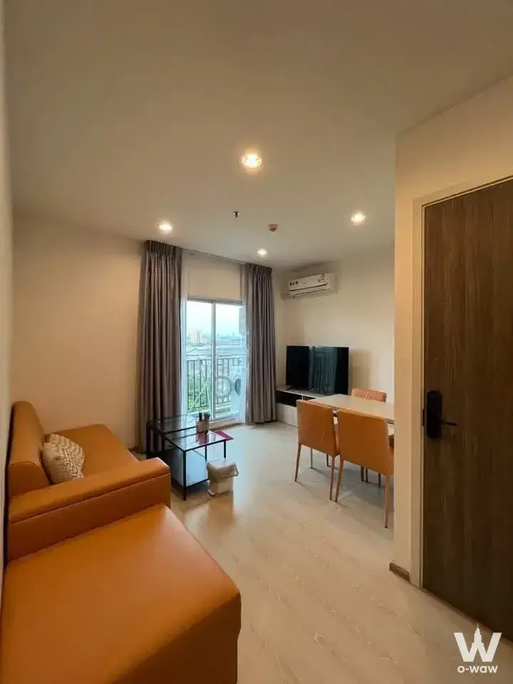 The living room of a 2-bed Niche Mono Sukhumvit Puchao condo has an orange-tan sectional and balcony view